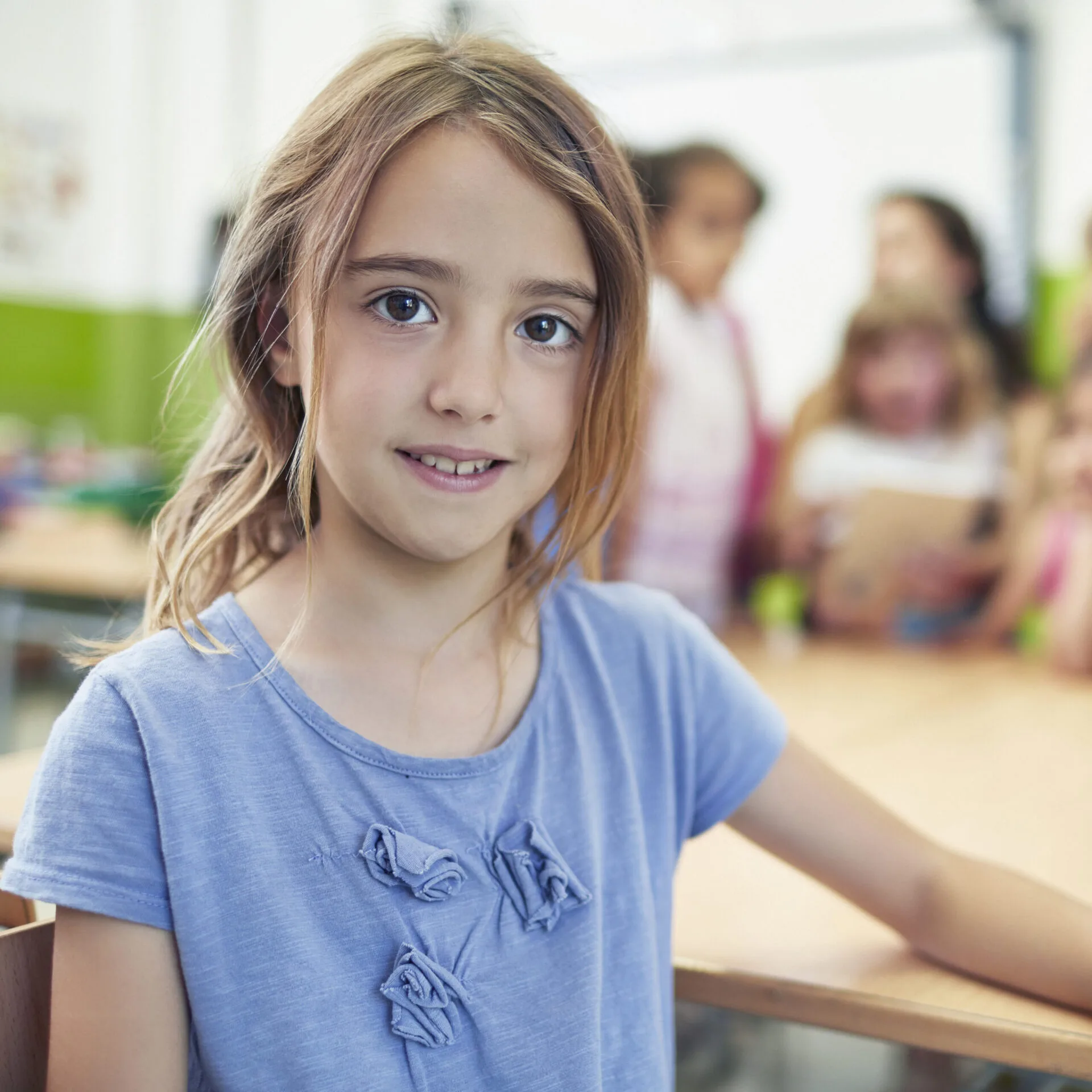 Cute smiling girl sitting at desk. Female student is with classmates in background. She is wearing blue casuals.
